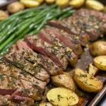 Juicy steak with herbs, roasted baby potatoes, and green beans arranged on a baking sheet.