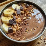 A close-up view of a chocolate peanut butter smoothie bowl topped with banana slices and peanuts.