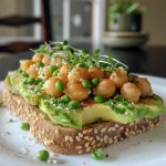 ['Close-up view of a High-Protein Chickpea Avocado Toast topped with microgreens and chili flakes.', 'Side view of a colorful chickpea avocado toast garnished with hemp seeds and thin tofu slices.', 'Delicious chickpea and avocado toast on whole-grain bread, featuring a sprinkle of chia seeds.', 'High-protein toast with mashed chickpeas, avocado, and vibrant greens, presented on a wooden board.']