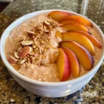 ['A close-up view of a Peach Smoothie Bowl topped with fresh peach slices, granola, and almonds.', 'A vibrant Peach Smoothie Bowl displayed from the side, featuring juicy peaches and crunchy toppings.', 'A detailed side shot of a Peach Smoothie Bowl, showcasing its creamy texture and colorful toppings.', 'A visually appealing Peach Smoothie Bowl with sliced peaches and granola, captured in natural light.']