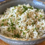 ['Close-up view of a bowl of fluffy white rice garnished with green parsley.', 'Side view of a serving of rice in a bowl with a sprinkle of garlic powder and butter.', 'Detailed close-up of cooked rice with fresh parsley, showcasing its texture.', 'Side perspective of a rice dish with moist grains and herbs on a wooden table.']